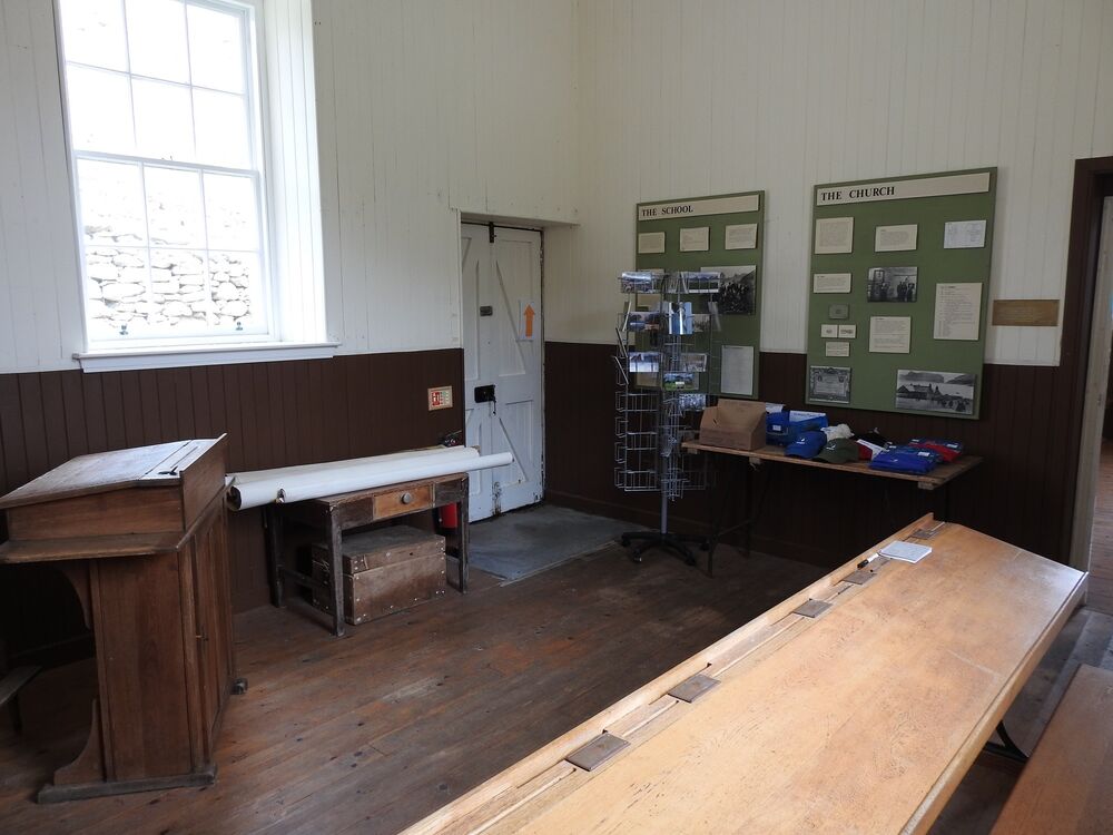 An old-fashioned school room has long wooden desks set around the edge of the room. At the far side, the desk is laid out with small souvenirs. Panels on the wall give information about how this room used to be used.