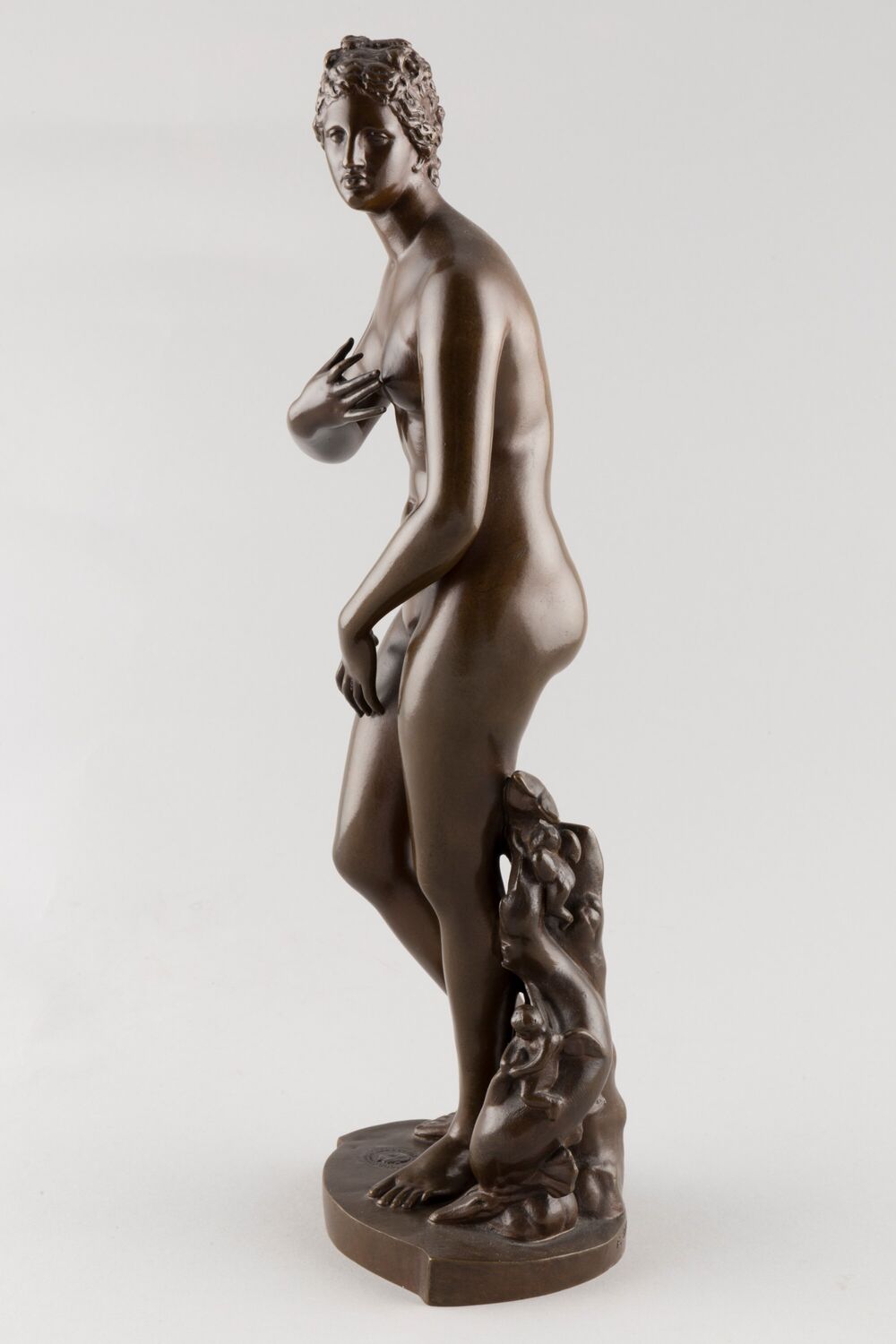 Side view of a bronze statue of Venus, with her right hand covering her left breast and her left hand on her right thigh.