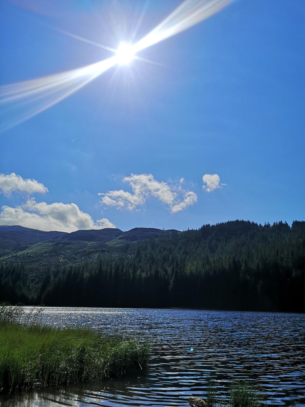 The sun shimmers on the ripples in a loch on a summer day. The sky and water are bright blue, with the rays gleaming in the sky. Mountains and woodland can be seen behind the loch.