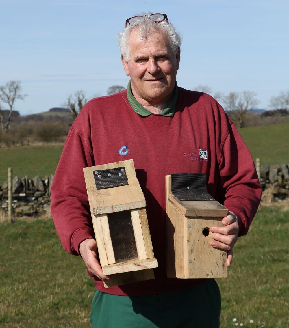 A man stands in a field holding a wooden squirrel feeder box in one hand and a wooden nest box in the other. He is wearing a bright red jumper and has his glasses on top of his head.