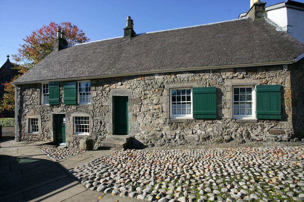 A view of the exterior of Weaver’s Cottage from the cobbled courtyard in front. It’s a stone single-storey cottage, with green wooden shutters beside the windows. There is an inscription on the lintel above the door.