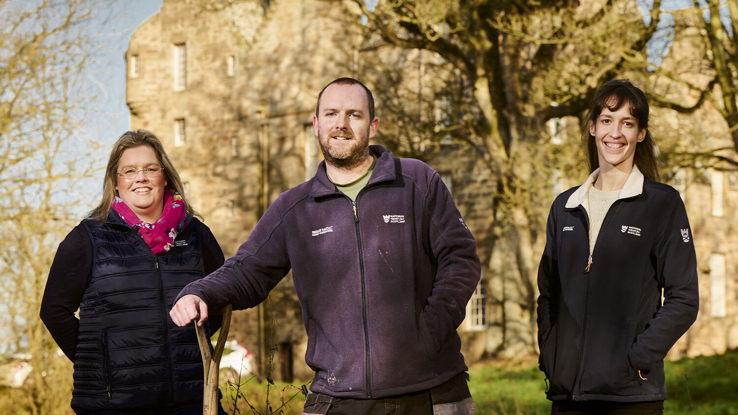 Three National Trust for Scotland staff members stand in front of Kellie Castle: a woman in a coat and a scarf, a man leaning on a gardening fork, and a woman in a Trust jacket.