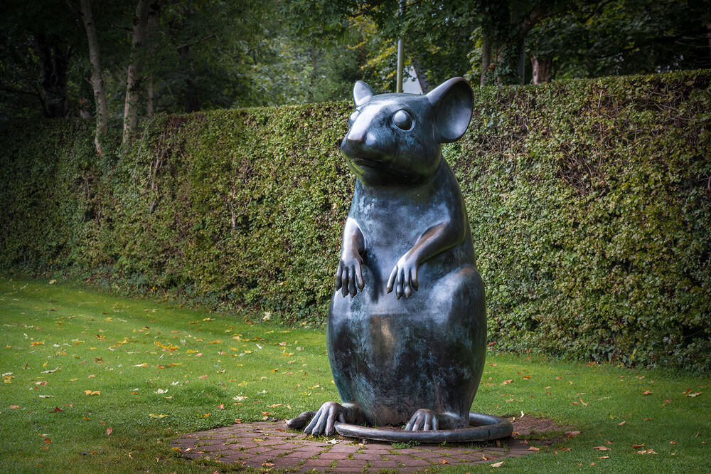 A human-sized statue of a mouse sat up with its paws in front of its body as it gazes into the distance. There is a tall hedge and trees behind it.