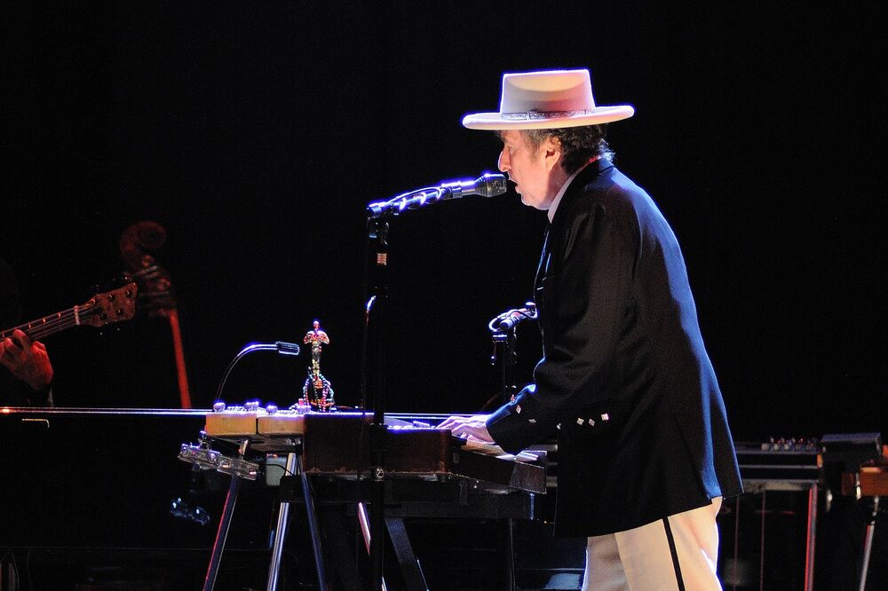 Bob Dylan, wearing a white panama hat and a dark jacket, stands behind a keyboard on stage, singing into a microphone.