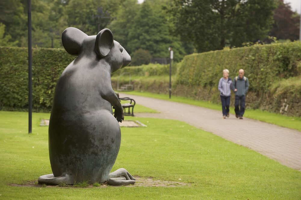 A very large stone statue of a mouse sitting on its hind legs stands on a patch of grass beside a long, straight path. In the background, a couple walk arm in arm along the path. A large hedge borders the path to the other side.
