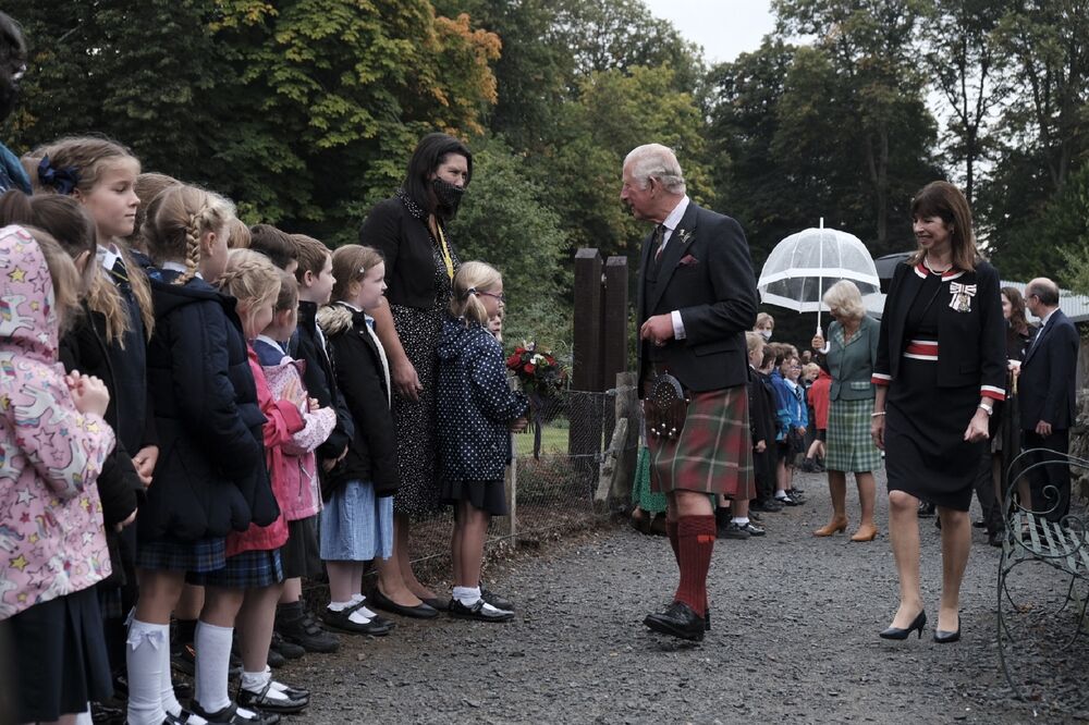 Prince Charles and other officials walk along a wide path, chatting to school children who line the sides. Prince Charles is wearing a kilt.
