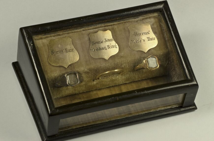 A small, wooden display box contains three gold rings in a row, all nestled in a cushion. Small gold plaques lie above each one labelling them (from left to right): Burns' Hair; Bonnie Jeans Wedding Ring; Burns' wife's hair. The ring in the middle is a narrow gold band, but the rings either side are more like signet rings.