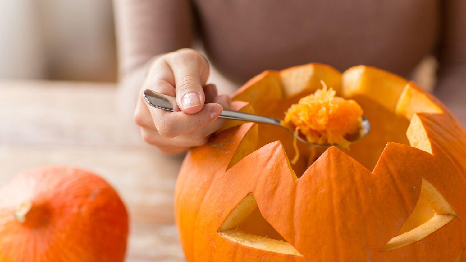 A lady scoops out flesh from a pumpkin that has been carved with a spooky face.