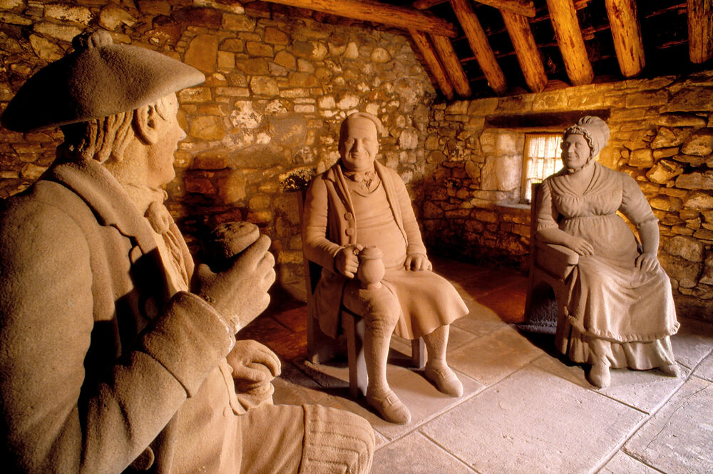 Carved sandstone sculptures are arranged in an outbuilding to depict a scene from the poem Tam o' Shanter, where Tam and his friends sit around a fireplace in a pub, drinking.