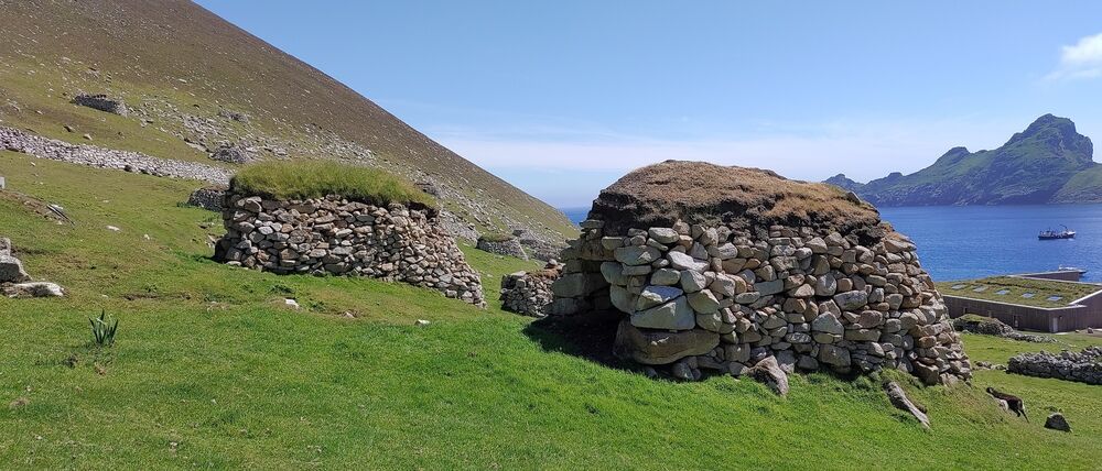 Two cleitean (stone structures with turf roofs) stand next to each other on a grassy hillside on a sunny day. The cleit in the foreground has a very dry turf roof; the one further back looks more grassy. The blue sea can be seen in the background.
