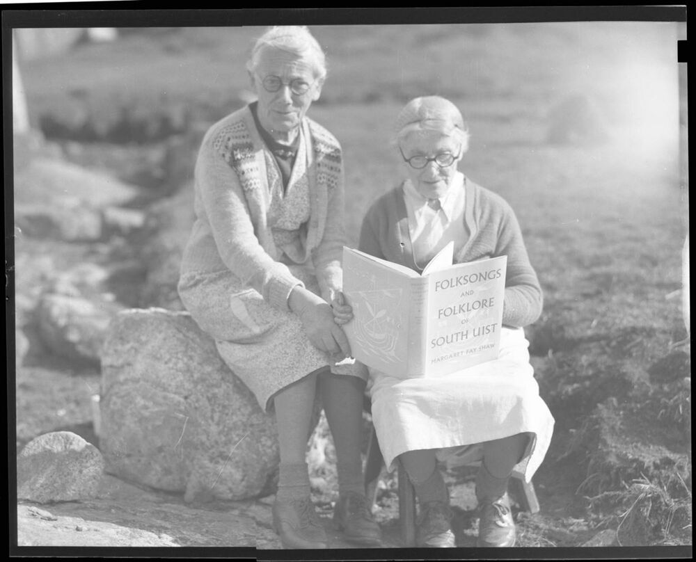 A black and white photo of two older women sitting on two large stones. Peigi MacRae sits to the right, holding a large book.