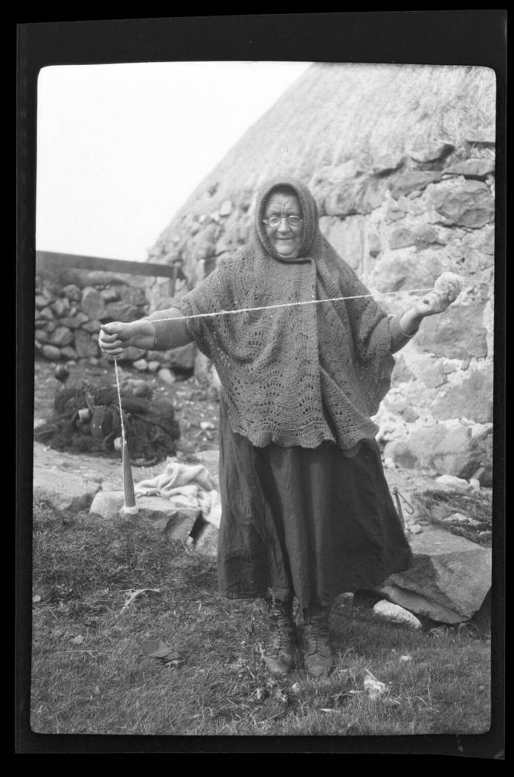 A black and white photo of an older woman spinning using a drop spindle. She stands outside wears a woollen headscarf.