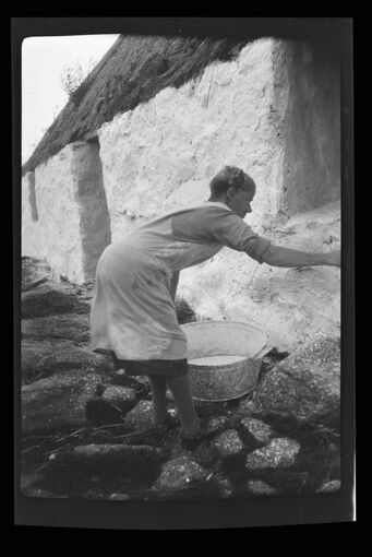 A black and white photo of an older woman whitewashing a stone cottage on South Uist.