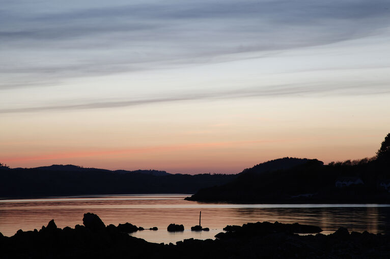 The sun sets over the coast at Rockcliffe, with the tree-covered hills silhouetted against a pink and purple sky.
