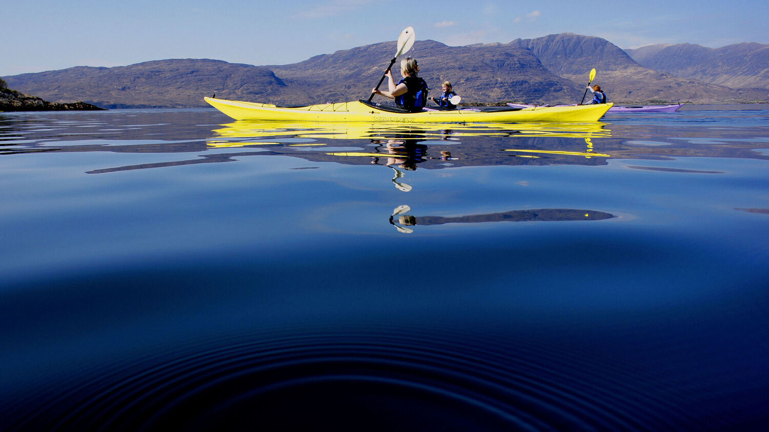A group of people in yellow kayaks paddle through still blue water. The image is taken from the surface of the water. In the distance the tall mountains of Kintail rise into a clear blue sky.