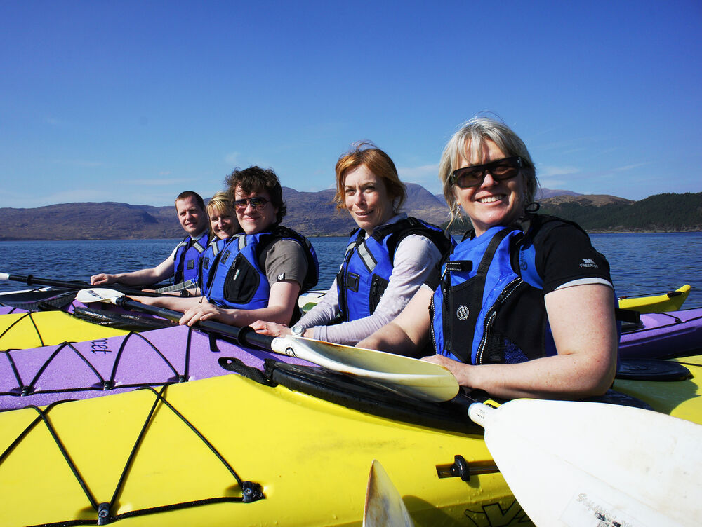 A group of five people line up in a row in their yellow kayaks out at sea. It is a glorious sunny day, and both the sky and water are a deep blue.