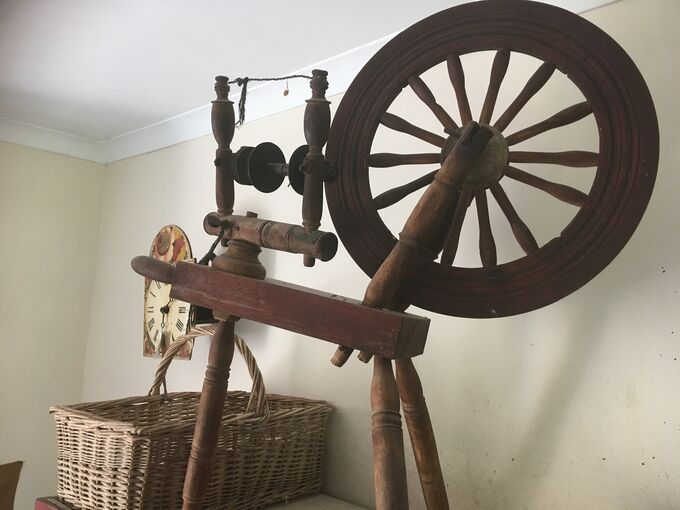 A wooden spinning wheel is displayed in a room, with a wicker basket in the background.