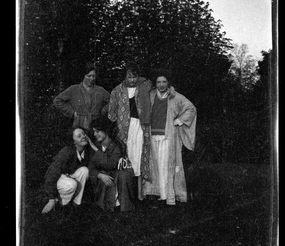 A black and white photo of five girls posing beside some trees. Three stand arm in arm, while the other two crouch on the ground before them. The girls wear dressing gowns and loose robes.