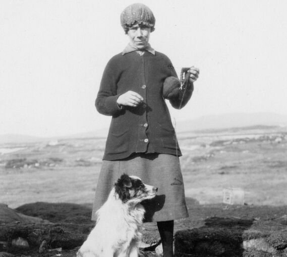 A black and white photo of a woman standing on a shore with a collie dog sitting at her feet. She wears a tight cloth hat, rather like a shower cap!