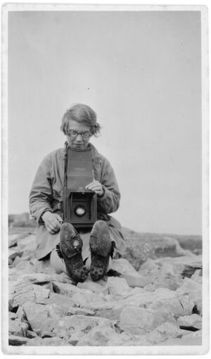 A young woman sits on a pile of rocks with her legs straight out in front of her. Around her neck she wears a large box camera with the shutter open.