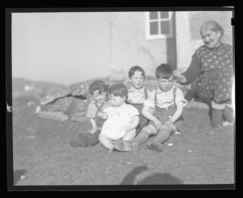 A black and white photograph of four very young children sitting together on some grass outside a croft house. An older woman sits just behind them, touching the ear of the closest boy. The shadow of the photographer can be seen on the lawn, with an arm raised in the air.