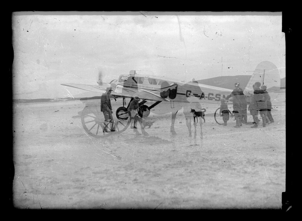 A black and white double exposed photograph. One layer shows a plane landing on a sandy beach with a group of people and a collie dog waiting nearby. The other, fainter image shows a horse and cart.