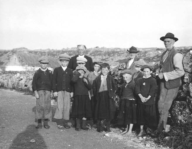 A black and white photograph of a group of young boys, with three older men standing behind them. They stand on a stone road, with some croft houses in the background. Some of the boys are barefoot. Most are scrunching up their faces, or shielding their eyes, against the sun. The shadow of the photographer falls to the left of the photo.