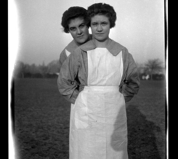 A black and white photograph of two women standing one in front of the other. Both wear white aprons and grey blouses. The woman at the front tucks her hands into the waistband of her apron.
