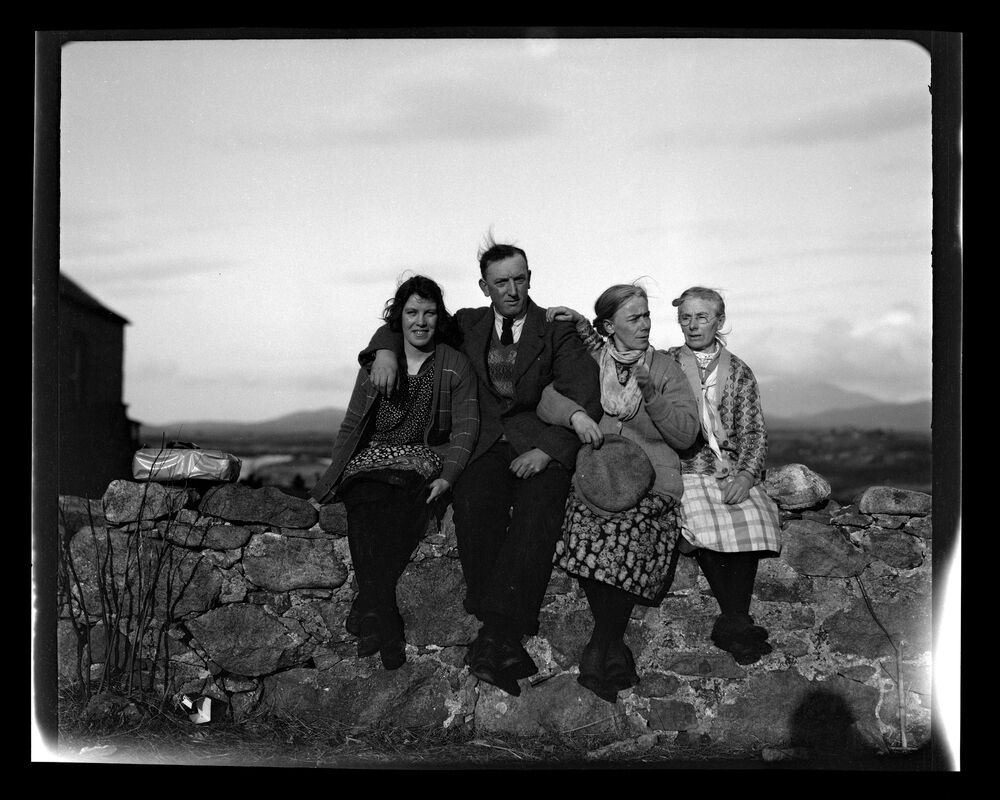 A black and white photograph of three women and a man sitting close together on a stone wall. All are well-wrapped up and have their arms around each other. The shadow of the photographer wearing a round hat can just be seen against the wall in the bottom right corner.