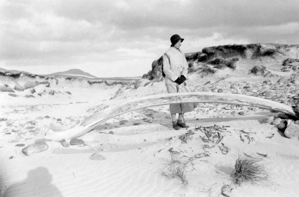 A black and white photograph of a woman standing on a sandy beach with dunes, just behind a very large jaw bone, probably from a whale. The shadow of a person wearing a hat is cast upon the sand in the bottom left corner.