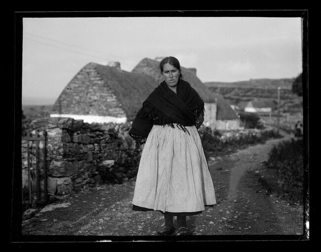 A black and white photograph of a woman walking along a stone road past croft houses. She wears a dark woollen shawl around her shoulders and has a paler skirt. The long shadow of the photographer falls along the road on the right.