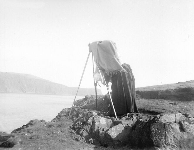 A woman has her head covered by a blanket as she adjusts her camera on a tripod, perched on some rocks by the edge of a cliff.