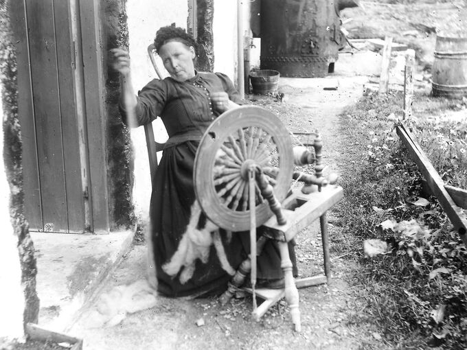 A woman sits at a spinning wheel holding some thread, just in front of a stone cottage. She wears a long black dress.