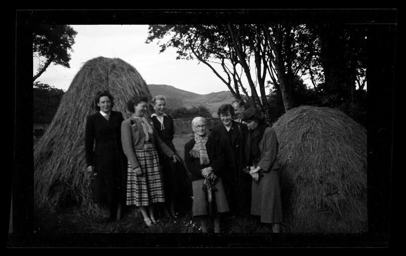 A black and white photograph of a group of women standing between two large hay stacks in a garden. An older lady stands in the middle, with the others surrounding her.