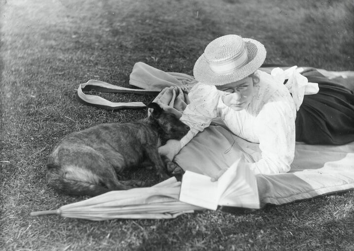 A lady wearing a white blouse and straw boater lies on a rug on the grass, her parasol beside her. She holds an open book in one hand. A sleeping dog rests its head on her arm.