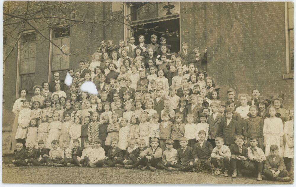 A sepia photograph of a large group of schoolchildren (over 100), standing or sitting on a set of steps outside a school building. Many of the girls wear white smocks or blouses; most boys wear a darker wool jacket. Teachers line the steps at the edge of the group. A tree branch with new buds is in the foreground.