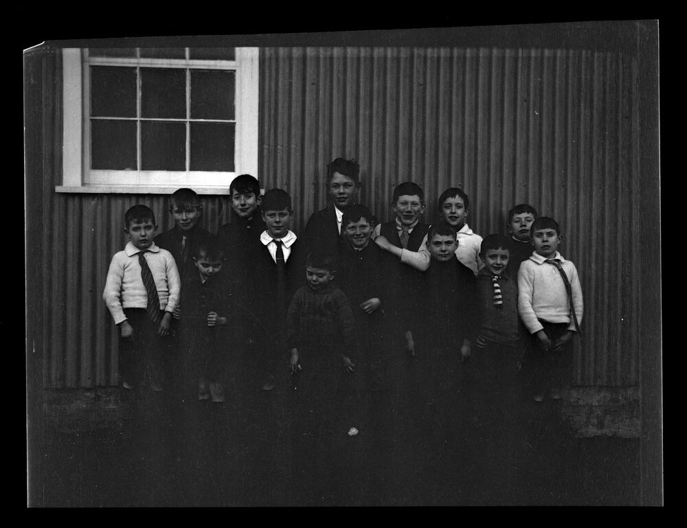 A black and white photograph of a group of 14 schoolboys (of mixed ages) standing in front of a corrugated schoolhouse and a white-framed window. Most of the boys are wearing a tie.