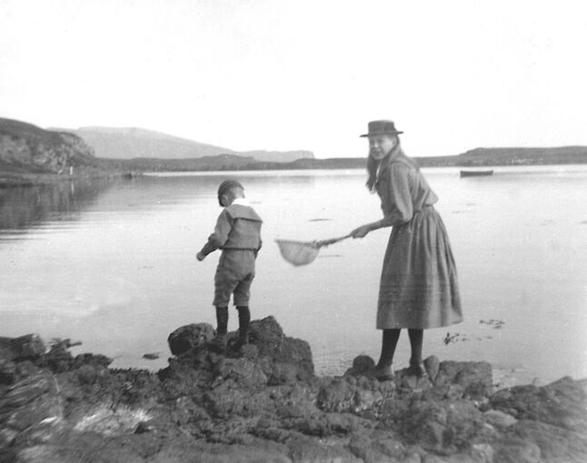 A young teenage girl and a young boy stand on rocks at the edge of the sea, holding a shrimping net. The water is very still and a small boat is anchored just off shore.