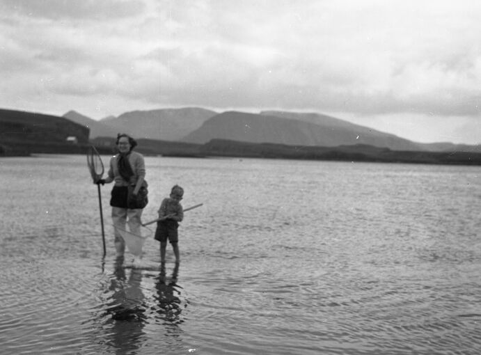 A woman and a young boy paddle in shallow water, both holding shrimping nets. Hills can be seen across the bay.