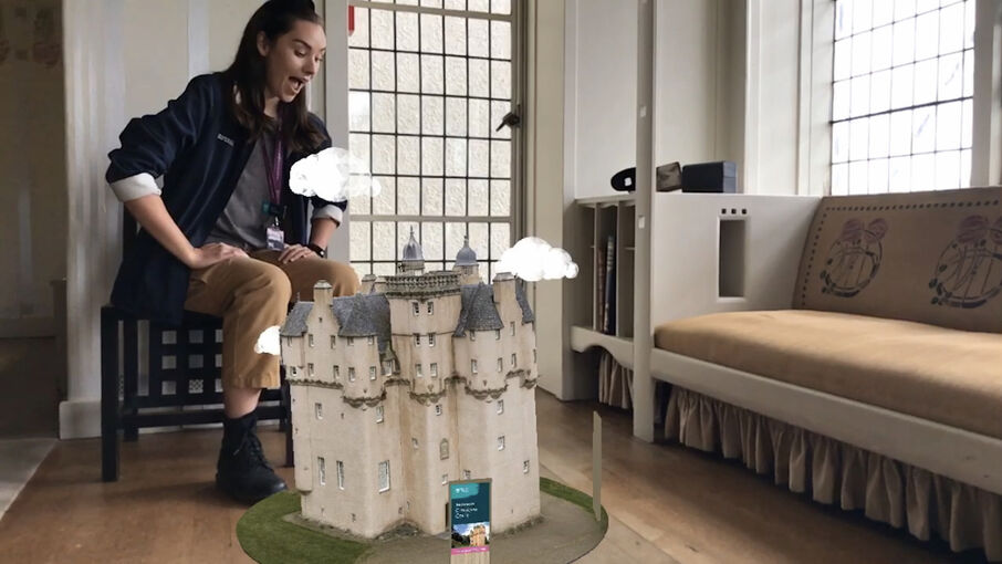 An augmented image of a castle appears on the wooden floor of the parlour at the Hill House. A woman sits on a Mackintosh-designed stool behind it, looking at it in wonder.