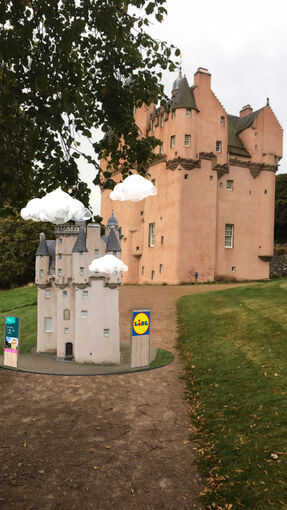 An augmented image of a castle appears on a gravel path, with the real castle in the background. The virtual image has white clouds bobbing over it, and the Lidl logo on a structure next to the castle.