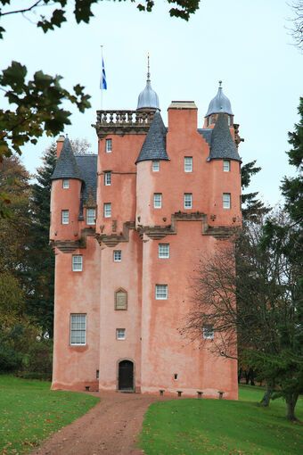 Front view of the pink-walled Craigievar Castle. A gravel path leads to the small front door, with trees at the side. A saltire flag flies from the roof.