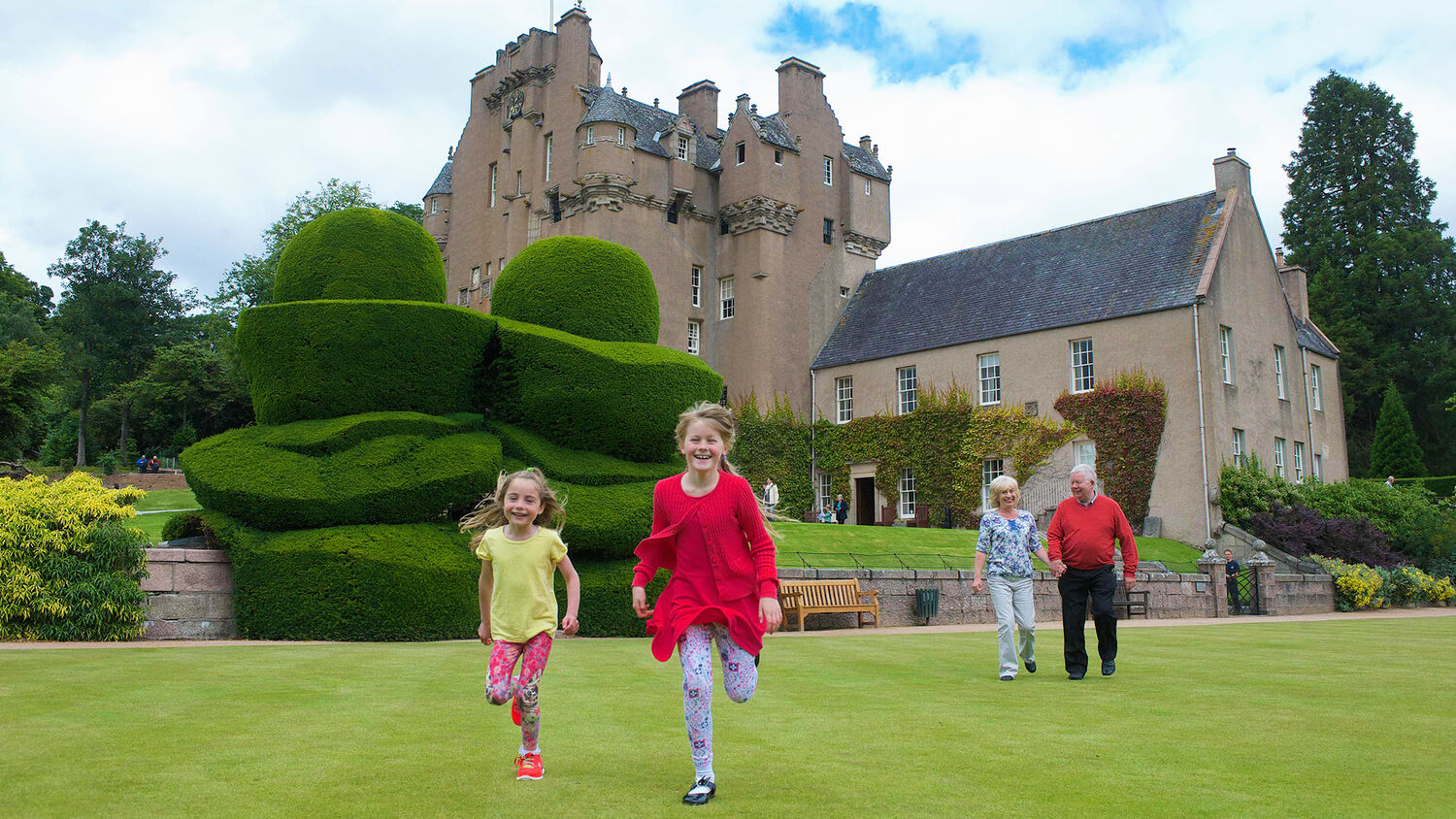 Tow children run towards the camera on the lawn in front of Crathes Castle. An older couple walk hand in hand behind them.