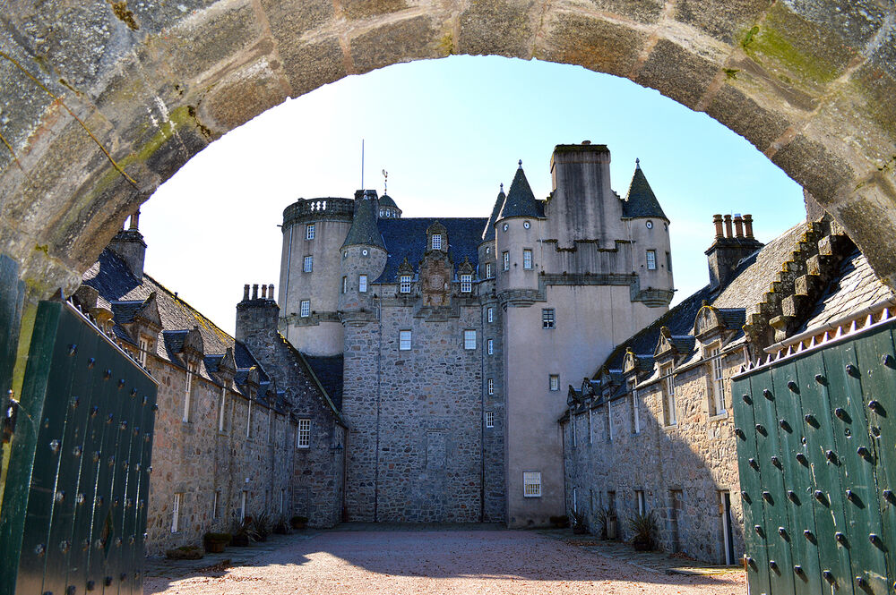 A view looking through the curved archway into Castle Fraser courtyard, with the castle at the far end. The courtyard appears to be gravelled with rows of outbuildings running either side.
