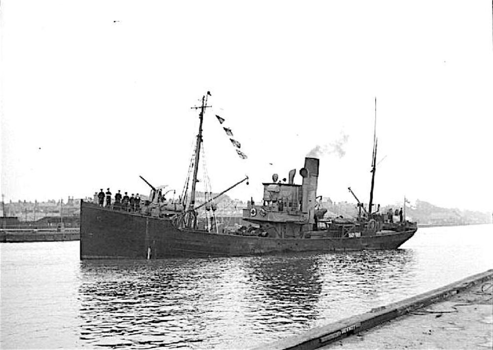 A black and white photograph of a steam trawler sailing along a river. Sailors stand on deck on the bow of the ship looking towards the dock. Flags are suspended from the mast line, and a funnel in the middle emits smoke. Large guns can be seen on deck at either end of the vessel.
