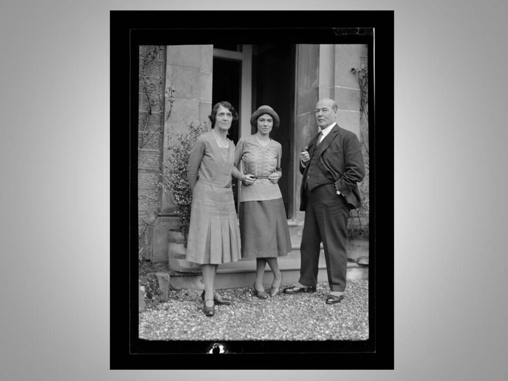 A black and white photo of three people standing in the doorway of a large, stone house. A young woman stands between her parents, who are dressed quite formally.