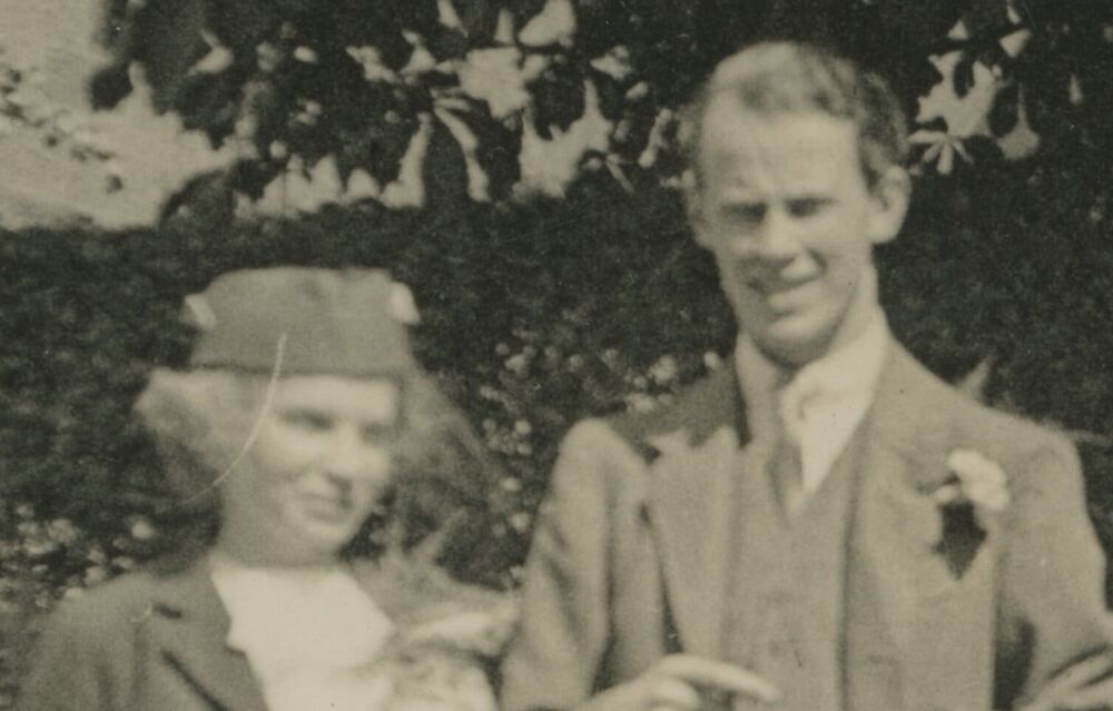 An old and slightly blurry black and white photo of a young couple on their wedding day in the 1930s. The man on the right wears a suit with a flower in the buttonhole. The woman on the left wears a small hat with a short veil from the brim.