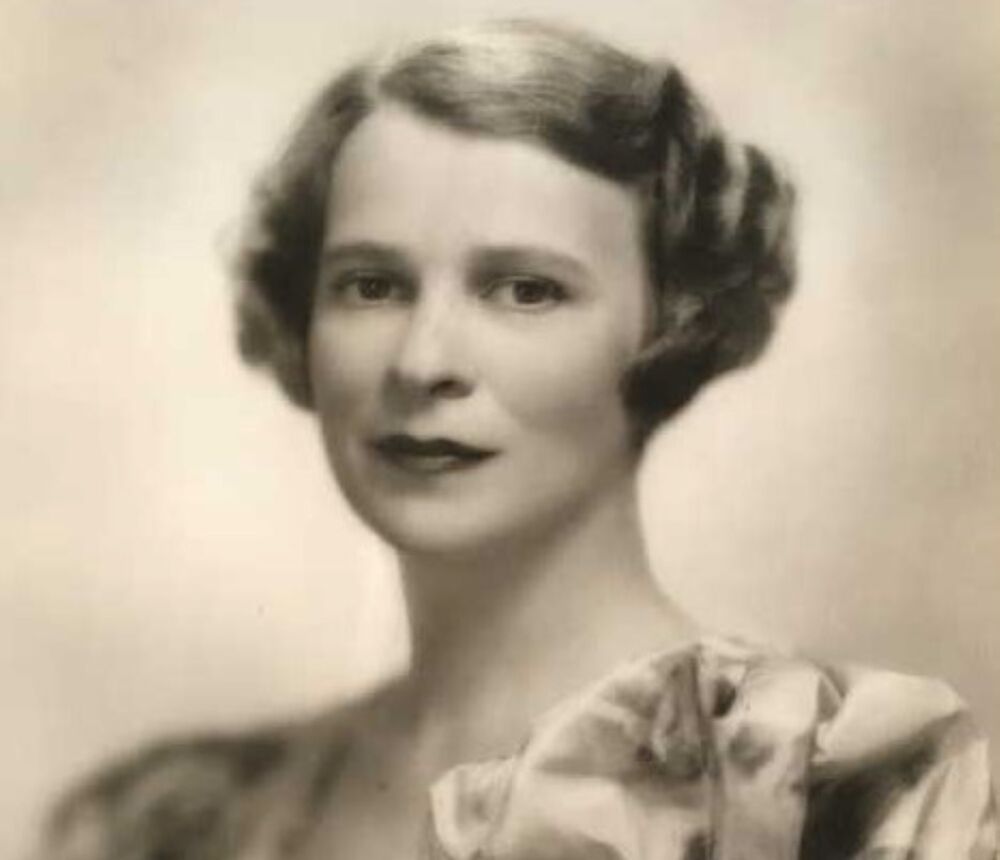A black and white portrait photograph of a young woman in the 1930s, showing her head and shoulders. She wears a floral satin dress and her short hair is waved.