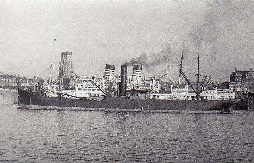 A black and white photograph of a long steam ship, with a large funnel in the centre emitting smoke. It is sailing close to a dockside.
