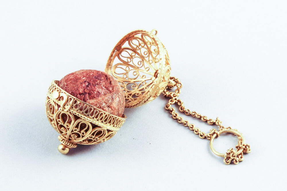 An ornate gold filigree case, containing a bold red bezoar. The case is opened wide and has a long gold chain